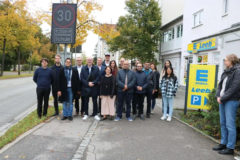 A group of Peregrine Technologies team members, partners from T-Systems, and local stakeholders stand together near a dynamic speed limit sign displaying '30' in a school zone in Landshut, Germany. The group is gathered on a sidewalk, surrounded by autumn trees and buildings, marking the successful launch of the 5-SAFE AI-powered traffic safety system to enhance school zone safety.